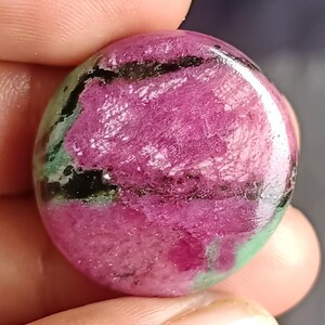 Op de afbeelding: Een gepolijste, ovaalvormige edelsteen met een levendige kleurenmix. De steen heeft een dominante magenta tint, afgewisseld met zwarte en groene insluitsels. Het oppervlak is glad en reflecterend, en toont de natuurlijke patronen in de edelsteen.