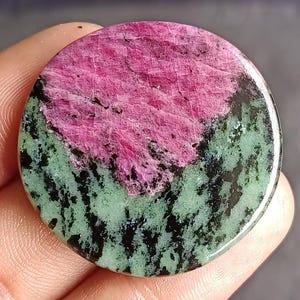 Könnte beinhalten: Ein runder Edelstein mit einem lebendigen Farbmuster. Die obere Hälfte ist ein tiefes Pink, das in eine hellgrüne und schwarz gesprenkelte Basis übergeht. Die polierte Oberfläche reflektiert das Licht und hebt die natürlichen Texturen hervor.