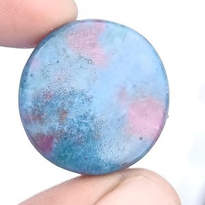 Op de afbeelding: Een ronde, doorschijnende edelsteen met een mix van blauwe, roze en turquoise kleuren. De steen heeft een gemarmerd uiterlijk met een gepolijst oppervlak. De edelsteen wordt tussen twee vingers gehouden.