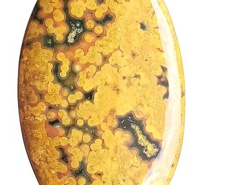 May include: An oval gemstone with a polished surface. The stone displays a pattern of yellow, black, and green spots and swirls. The gemstone is held between two fingers, highlighting its size and detail. A natural stone for jewellery making.