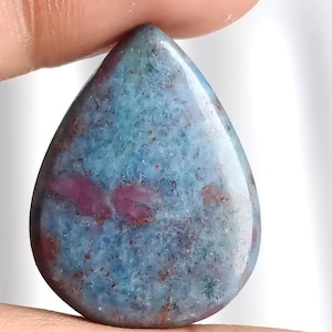 Puede incluir: Una gema pulida en forma de lágrima. La piedra presenta una mezcla de tonos azules, rojos y marrones, con un patrón moteado. La gema se sostiene entre dos dedos.