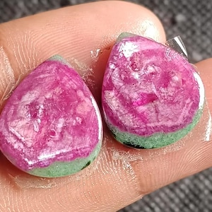 Op de afbeelding: Twee druppelvormige edelstenen. De edelstenen hebben een levendig roze centrum met een groene basis. De stenen worden in een hand gehouden, waarbij hun gepolijste oppervlakken en natuurlijke kleurvariaties worden getoond. Voor sieraden.