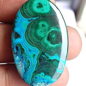 Può includere: Pietra preziosa ovale con un motivo sorprendente di verdi e blu vorticosi. La superficie lucida rivela cerchi concentrici e linee fluide, creando un design naturale unico. I colori della pietra variano dal verde acqua intenso al turchese vibrante, con aree di verde scuro.