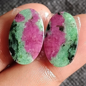 May include: Two oval-shaped ruby zoisite gemstones. The gemstones feature a vibrant mix of colors, with a prominent magenta hue contrasted by areas of green and black. The stones are held in place by a clear adhesive.