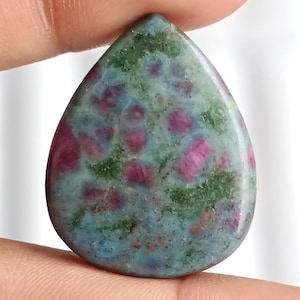 May include: A teardrop-shaped gemstone with a mottled pattern of deep red, green, and blue hues. The polished surface reveals a mix of colours, with the red inclusions adding contrast. The stone is held between two fingers.