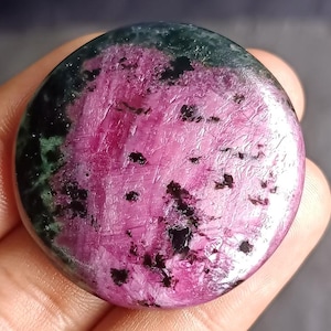 Può includere: Una gemma rotonda e lucidata con una vibrante tavolozza di colori. La pietra presenta un mix di rosa intenso, nero e verde scuro. La superficie ha un aspetto strutturato con motivi e inclusioni visibili, creando un'estetica unica e naturale.