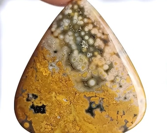 May include: A polished, triangular gemstone with a distinctive pattern. The stone displays a mix of colours, including mustard yellow, cream, black, and grey. The surface has a speckled and swirling design, with a glossy finish. The gemstone is held between two fingers.