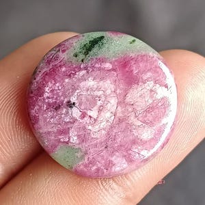 Può includere: Una gemma rotonda con una superficie lucida. La pietra presenta una miscela di colori, tra cui rosa, verde e bianco. Il rosa domina il centro, mentre accenti verdi e bianchi sono visibili nella parte superiore. La pietra è tenuta in una mano.