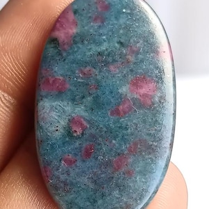 Può includere: Pietra preziosa ovale con superficie lucida. La pietra presenta un colore di base verde acqua con macchie irregolari di rosa intenso e piccole inclusioni nere. La gemma è tenuta in una mano.