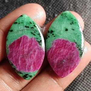 Op de afbeelding: Twee ovale edelstenen. Elke steen heeft een basis van felroze robijn, bedekt met een groene laag met zwarte insluitsels. De stenen zijn gepolijst en hebben een glad, reflecterend oppervlak dat natuurlijke patronen laat zien.