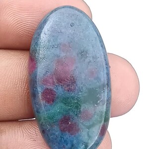 Puede incluir: Gema ovalada con una mezcla de colores. La piedra presenta una base de tonos azules y verdes, con manchas dispersas de rojo intenso. La superficie pulida refleja la luz, resaltando los patrones y texturas naturales.