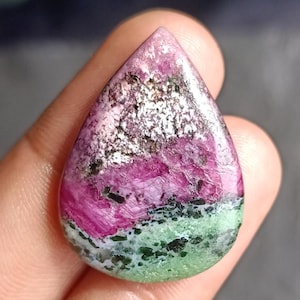 Peut inclure: Une pierre précieuse en forme de goutte polie. La pierre présente des couches de rose, de blanc, de vert et de noir. La pierre est tenue dans une main, soulignant sa taille et sa forme. Les couleurs et les motifs de la pierre créent un aspect unique. Mesure environ 3 cm de long.