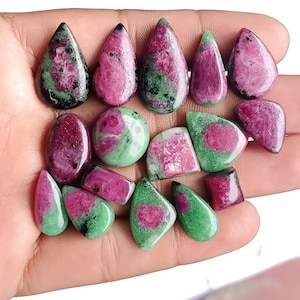 Op de afbeelding: Een verzameling gepolijste robijn in zoisiet edelstenen in verschillende vormen, waaronder druppels, ovalen en onregelmatige vormen. De stenen vertonen een mix van diepe roze, groene en zwarte kleuren. De edelstenen worden in een hand gehouden.