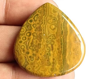 Puede incluir: Una gema pulida en forma de lágrima con un patrón único. La piedra presenta una mezcla de tonos amarillos, marrones y verdes, con patrones circulares y lineales. La superficie es lisa y brillante, mostrando la belleza natural de la piedra.