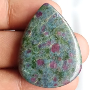 May include: A teardrop-shaped gemstone with a polished surface. The stone features a mottled pattern of green, blue-gray, and deep red spots. The gem is held in a hand, showcasing its size and shape.