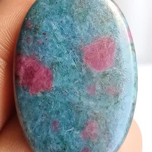 Puede incluir: Gema ovalada con base turquesa e inclusiones de rubí. La piedra pulida muestra una mezcla de texturas y colores, creando una apariencia única. Ideal para joyería o coleccionismo. Medidas en centímetros.