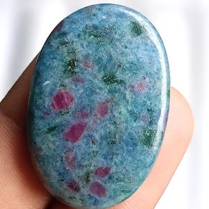 Può includere: Pietra preziosa ovale con superficie lucida. La pietra presenta una miscela di tonalità blu, verdi e rosse. Le inclusioni rosse sono sparse, creando un motivo unico. La gemma è tenuta in una mano.