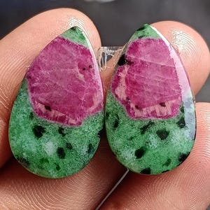 May include: Two teardrop-shaped ruby zoisite gemstones. The gemstones feature a vibrant pink center surrounded by a green base with black speckles. The stones are polished and have a glossy finish, perfect for jewelry making.