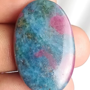 Könnte beinhalten: Oval geformter Edelstein mit polierter Oberfläche. Der Stein weist eine Mischung aus Farben auf, darunter Türkis, Blau, Grün und Rosa. Der Edelstein wird zwischen den Fingern gehalten und zeigt seine natürliche Schönheit und einzigartigen Farbvariationen.