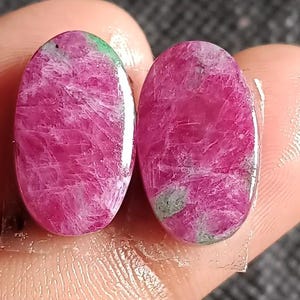 Könnte beinhalten: Zwei ovale Edelsteine in leuchtendem Rosa, die eine marmorierte Textur mit Streifen von hellerem Rosa und Hauch von Grün aufweisen. Die Edelsteine werden in einer Hand gehalten, mit einem transparenten Klebestreifen.