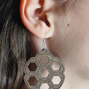 Puede incluir: Pendientes geométricos de madera con diseño de panal. Los pendientes son de color marrón claro y cuelgan de ganchos plateados. Los pendientes se llevan en la oreja de una persona.