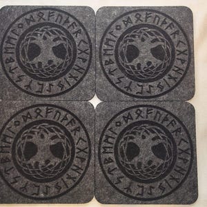 Op de afbeelding: Vier vierkante grijze onderzetters met een donkergrijs cirkelvormig ontwerp met een boom en runentekst. De onderzetters zijn gemaakt van een viltachtig materiaal en zijn vierkant gerangschikt.