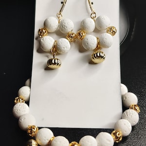 Könnte beinhalten: Ein Schmuckset mit weißen, porösen Steinperlen und goldfarbenen Akzenten. Das Set beinhaltet ein Armband und ein Paar Ohrringe. Die Ohrringe haben einen Hakenverschluss und einen kleinen, goldfarbenen Anhänger. Das Armband hat einen goldfarbenen Verschluss.