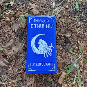 May include: A blue rectangular object with white text and a graphic of a creature with tentacles and a crescent moon. The text reads "THE CALL OF CTHULHU" and "H.P. LOVECRAFT". The object is resting on a bed of leaves and twigs.
