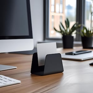 May include: A black metal business card holder with a minimalist design. A white business card is displayed in the holder. The holder is on a wooden desk, with a computer monitor and plants in the background.