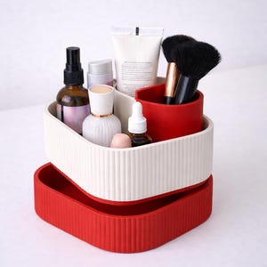Puede incluir: Un organizador de cosm&eacute;ticos de dos niveles, acanalado, en rojo y blanco roto. El nivel superior contiene varios productos de belleza, incluyendo botellas, tubos y brochas. El nivel inferior es una bandeja roja s&oacute;lida.