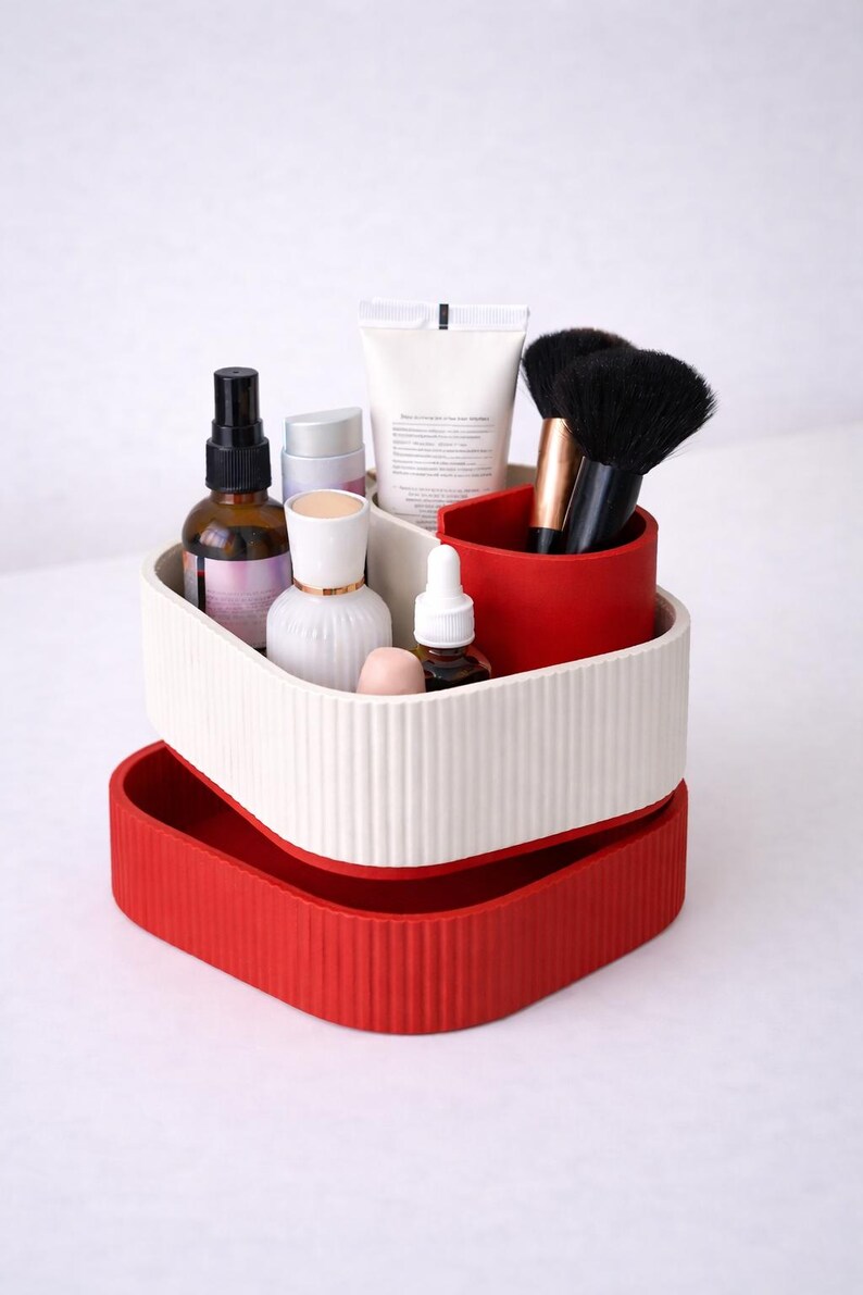 Puede incluir: Un organizador de cosm&eacute;ticos de dos niveles, acanalado, en rojo y blanco roto. El nivel superior contiene varios productos de belleza, incluyendo botellas, tubos y brochas. El nivel inferior es una bandeja roja s&oacute;lida.