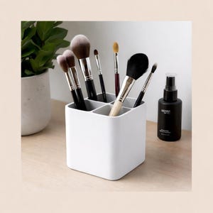 Puede incluir: Un soporte blanco cuadrado para brochas de maquillaje con varias brochas de maquillaje de diferentes tamaños y formas. A la derecha del soporte hay un frasco rociador negro con texto blanco. Una planta verde en una maceta blanca está en el fondo.