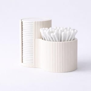 Puede incluir: Un organizador de baño crema de dos partes. Una sección contiene una pila de discos de algodón blancos, mientras que la otra contiene un paquete de bastoncillos de algodón blancos. Ambas secciones tienen detalles acanalados verticales, creando una estética limpia y moderna.