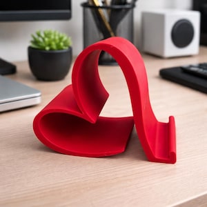 Puede incluir: Un soporte para teléfono rojo vibrante con forma de corazón, hecho de un material liso y curvado. El soporte está diseñado para sostener un teléfono en posición vertical. El fondo incluye un escritorio con útiles de oficina y una pequeña planta en maceta.
