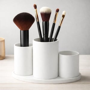 Puede incluir: Un soporte blanco de cerámica para brochas de maquillaje con tres compartimentos cilíndricos sobre una base ovalada a juego. El soporte contiene varios pinceles de maquillaje con mangos negros y cerdas marrones y blancas. El soporte está sobre una superficie clara.