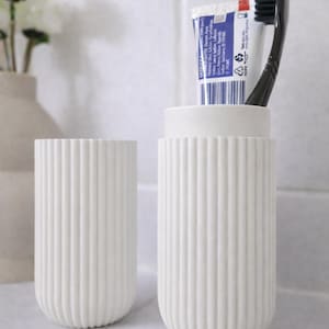 Pode incluir: Dois acessórios de casa de banho em cerâmica branca canelada. Um contém um tubo de pasta de dentes e uma escova de dentes preta. O outro está vazio. Os itens estão em uma superfície branca, sugerindo uma decoração de casa de banho moderna.