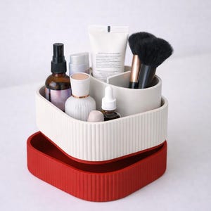 Puede incluir: Un organizador de dos niveles en blanco y rojo, lleno de productos de belleza. La secci&oacute;n superior es blanca con ranuras verticales y contiene botellas, tubos y brochas. La parte inferior es una bandeja roja s&oacute;lida.