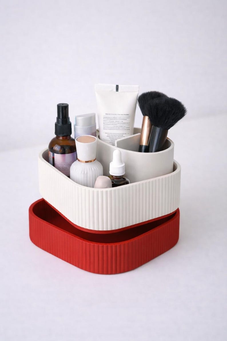Puede incluir: Un organizador de dos niveles en blanco y rojo, lleno de productos de belleza. La secci&oacute;n superior es blanca con ranuras verticales y contiene botellas, tubos y brochas. La parte inferior es una bandeja roja s&oacute;lida.