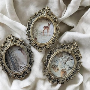 Peut inclure: Trois cadres ovales de style ancien avec des détails dorés ornés. Chaque cadre contient une petite illustration détaillée d'animaux de la forêt, dont un cerf et des lapins, sur un fond hivernal. Les cadres reposent sur un tissu blanc doux.