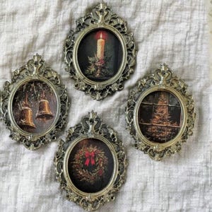 May include: Four vintage Christmas ornaments in ornate silver-coloured frames. Each features a different festive scene: a lit candle, bells, a wreath, and a Christmas tree. The ornaments are displayed on a neutral-coloured fabric.
