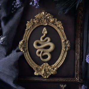 May include: Ornate gold-colored oval frame with a detailed snake emblem on a dark background. The frame is surrounded by dark fabric and sprigs of purple flowers, creating a gothic or mystical aesthetic.