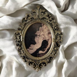 Può includere: Una cornice ovale in stile antico con dettagli dorati ornati. La cornice circonda un dipinto ovale scuro con una figura bianca simile a un uccello o un angelo. La cornice poggia su un tessuto bianco strutturato.