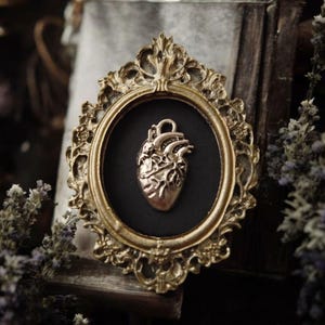 Puede incluir: Un marco dorado ornamentado, de forma ovalada, exhibe un colgante de corazón metálico detallado sobre un fondo negro. El conjunto se presenta sobre un fondo borroso con flores secas y un libro antiguo, creando una estética gótica.