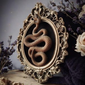 May include: A detailed, bronze-colored snake sculpture coiled within an ornate, gold-toned frame. The frame has intricate scrollwork and is set against a dark background. Dried lavender and white flowers are in the background, creating a gothic aesthetic.