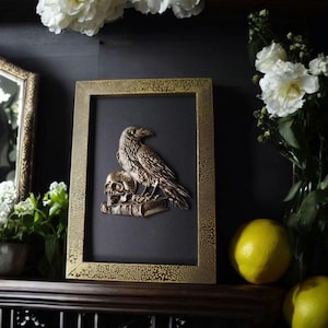 Puede incluir: Escultura metálica enmarcada de un cuervo posado sobre una calavera y libros. La obra de arte está sobre un fondo negro dentro de un marco dorado. La imagen está rodeada de flores blancas y limones.