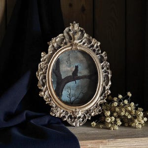 Puede incluir: Un marco ovalado de color dorado antiguo con una pintura de un gato negro posado en la rama de un árbol. La pintura tiene un fondo azul con ramas desnudas. El marco ornamentado se encuentra sobre una superficie de madera.