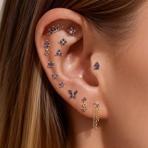 May include: An ear adorned with multiple gold earrings featuring various designs. The earrings include teardrops, butterflies, and floral shapes, all embellished with purple and blue gemstones. The jewelry is set against the backdrop of a person's ear.