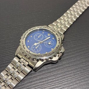 May include: A silver wristwatch with a blue face and multiple sub-dials. The watch has a stainless steel strap and a bezel marked with international city names. The watch is displayed on a dark surface.