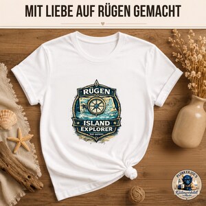 Könnte beinhalten: Ein weißes T-Shirt mit kurzen Ärmeln und einem zentralen Motiv, das einen Kompass über einer Landkarte zeigt, mit dem Text RÜGEN ISLAND EXPLORER EST OSTEES. Oben im Bild steht der Text MIT LIEBE AUF RÜGEN GEMACHT in einer dunklen Schrift.