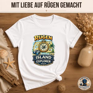 Könnte beinhalten: Ein weißes T-Shirt mit einem Grafikdruck aus Kompass, Fernglas und Palmen sowie dem Text RÜGEN ISLAND EXPLORER RÜGEN. Oben im Bild steht der Text MIT LIEBE AUF RÜGEN GEMACHT und in der Ecke befindet sich ein kleines Logo.